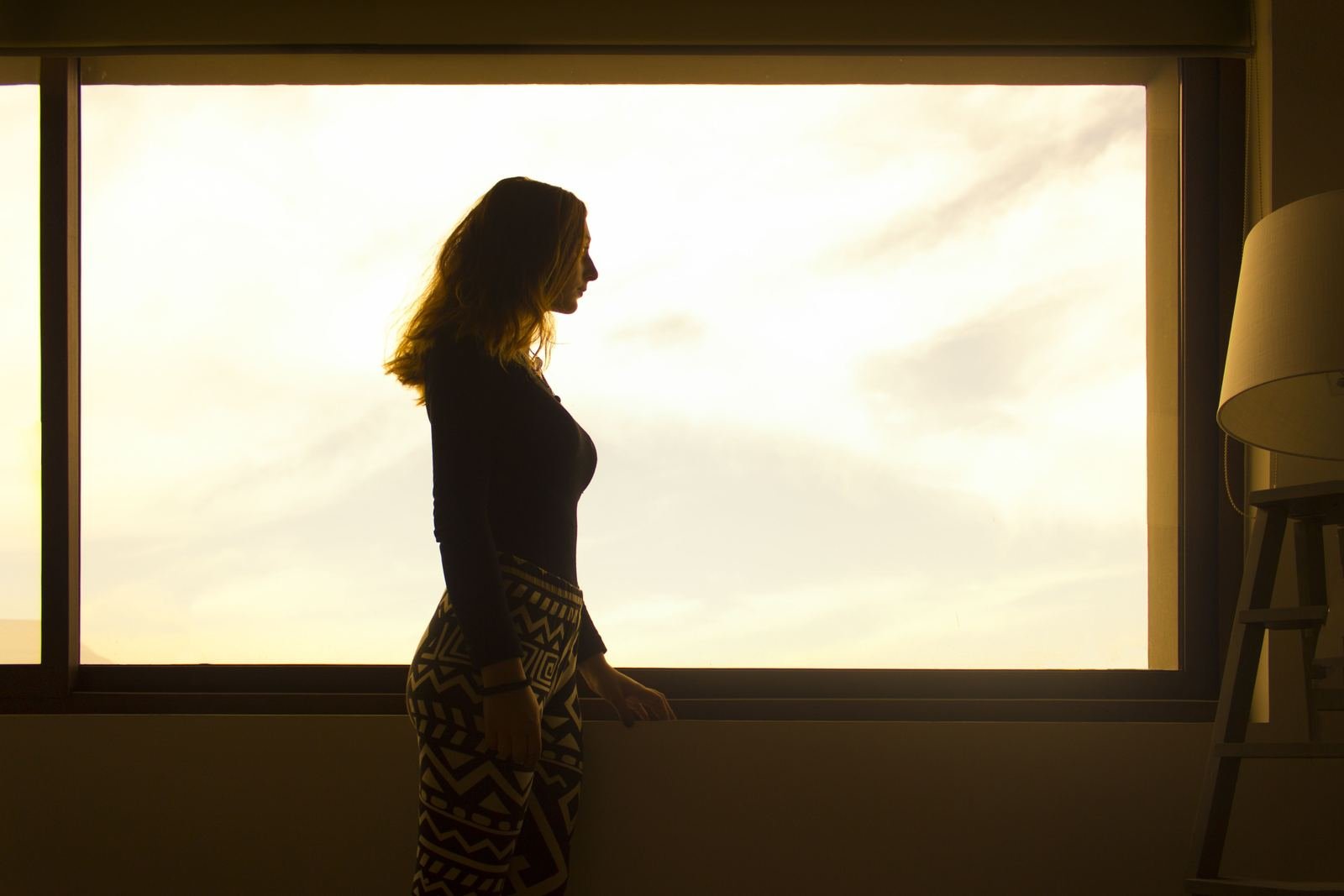 Silueta de una mujer junto a una ventana con luz cálida al atardecer, en un momento de reflexión